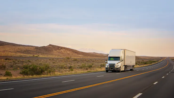 A white semi-truck pulling a white trailer drives down a highway, with hills in the background.