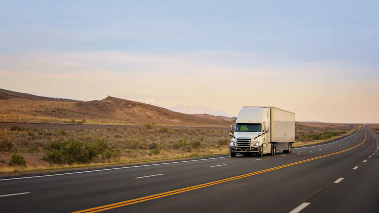 A white semi-truck pulling a white trailer drives down a highway, with hills in the background.
