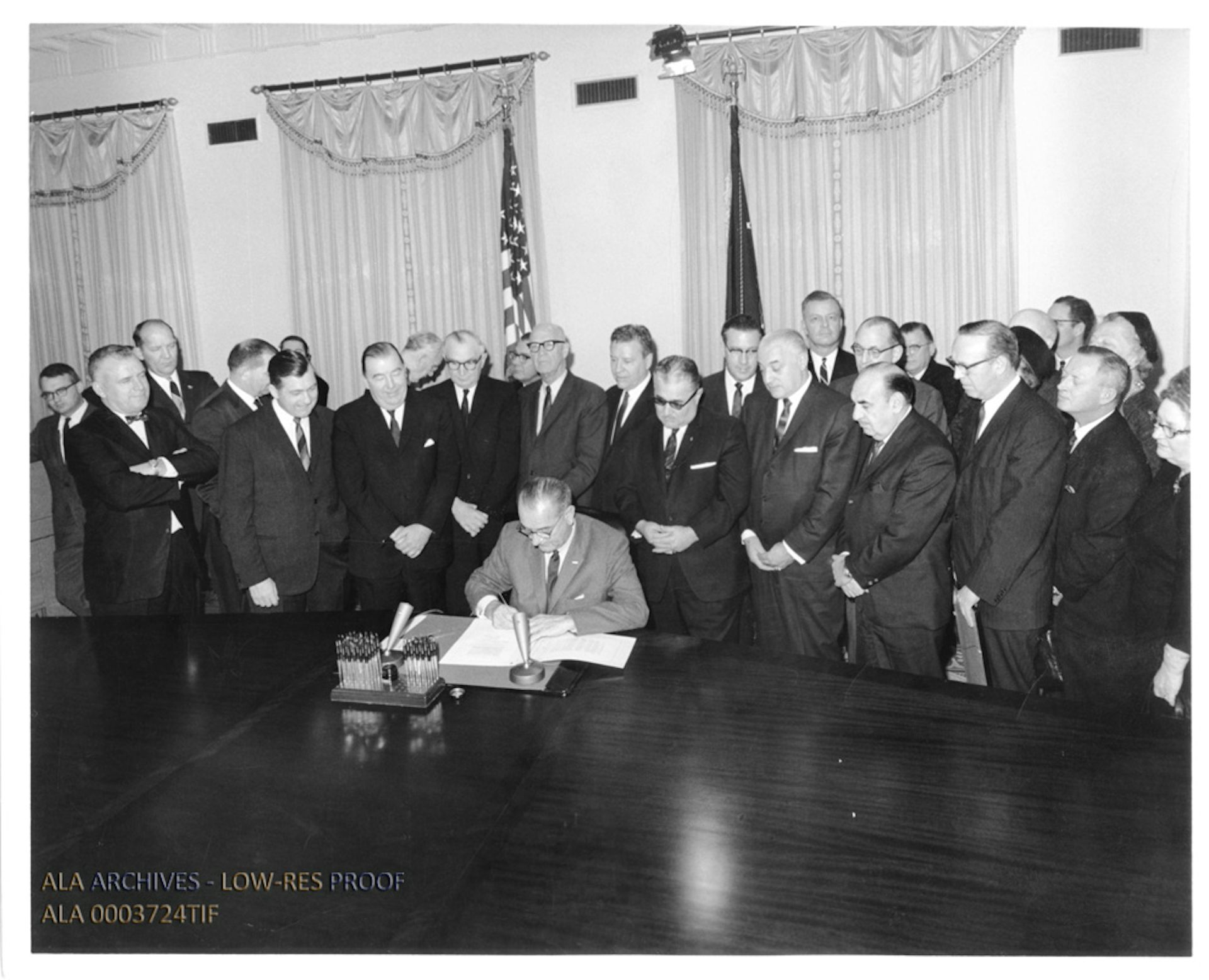 A black-and-white photo of a man seated at a desk signing a document as a crowd of people in suits stands behind him.