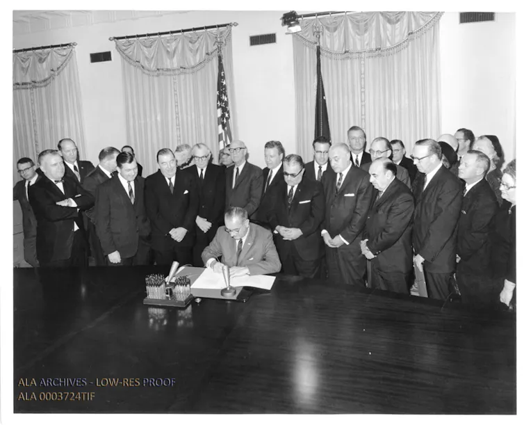 A black-and-white photo of a man seated at a desk signing a document as a crowd of people in suits stands behind him.