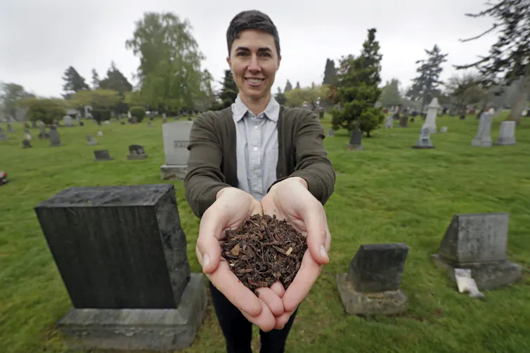 A woman with short hair posing in a cemetery holding a handful of mulch to the camera.