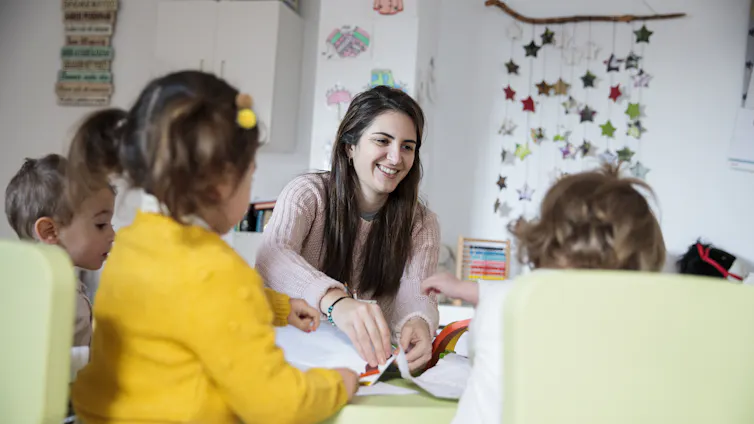 A female early childcare educator running a crafting activity with young children.