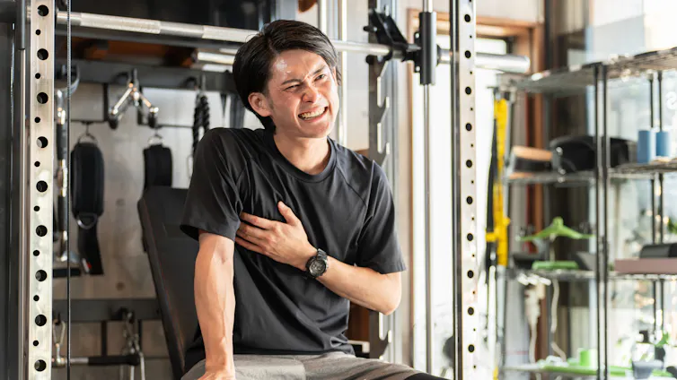 A young man using an exercise machine in the gym holds his chest while grimacing in pain.