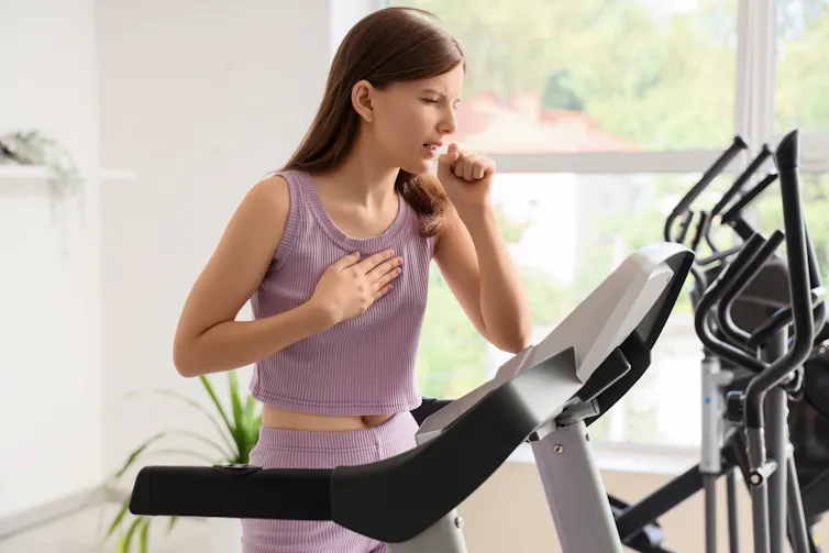 A young woman using a treadmill coughs while holding her chest in pain.