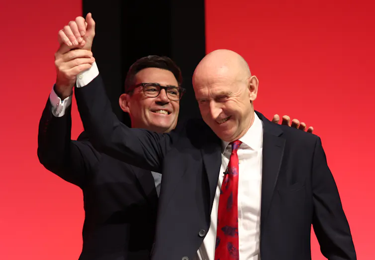 andy burnham holding the hand of chancellor john healey aloft at the labour party conference 2026.