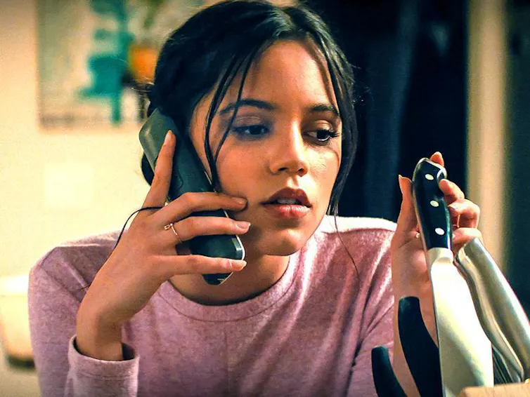 A woman holds a phone to her hear and in her other hand pulls a knife out of its holder.