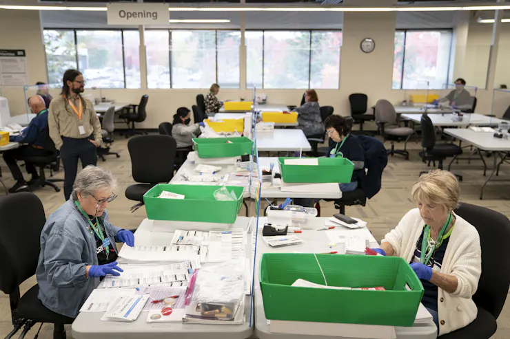 People working at a long table covered with papers and green boxes.