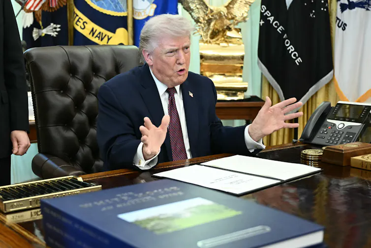 A man at a desk, gesturing with his hands.