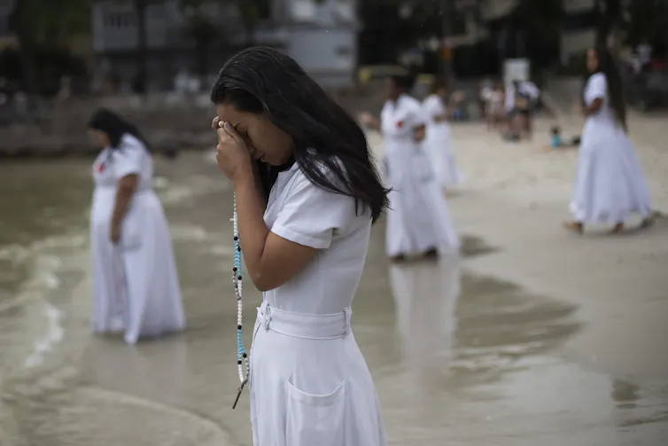 Several women in long white skirts and T-shirts stand in sand at the edge of the ocean.