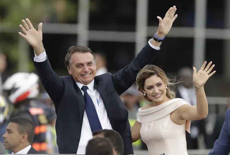 A man in a suit and a woman in a dress wave to a crowd outdoors.