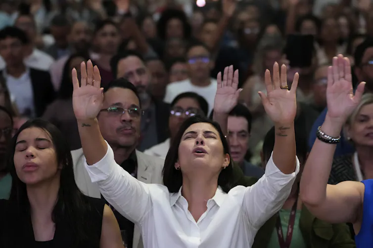 A woman in a white shirt, whose eyes are closed, holds her hands up in prayer as she stands amid many other people.