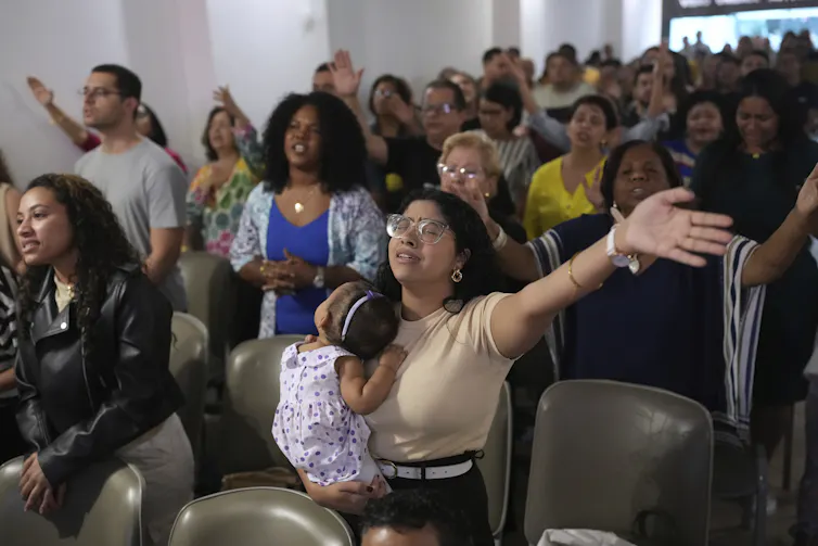 People standing as they sing or pray in a room with white walls and rows of chairs.