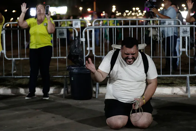 A man in a white T-shirt and shorts kneels on the street in prayer, with a praying woman standing near him.