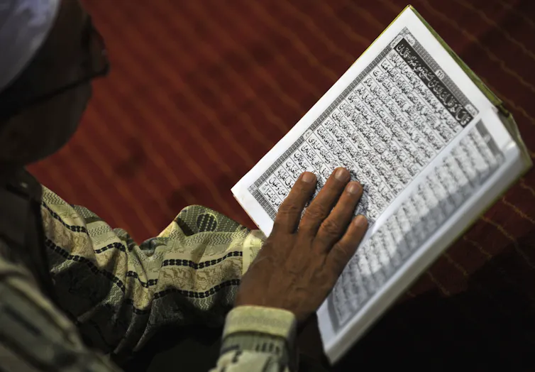 A woman with her head covered reads from a book written in Arabic.