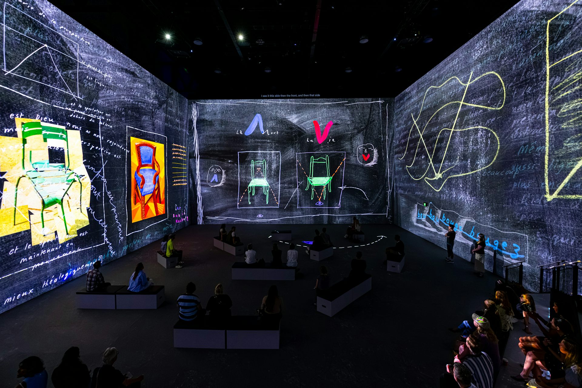 A large room with digital projections on the walls showing sketches. People sit on benches in the dark.