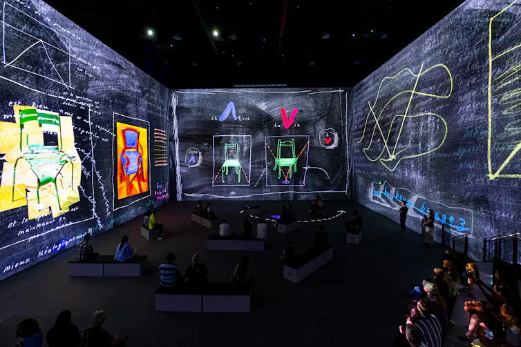 A large room with digital projections on the walls showing sketches. People sit on benches in the dark.