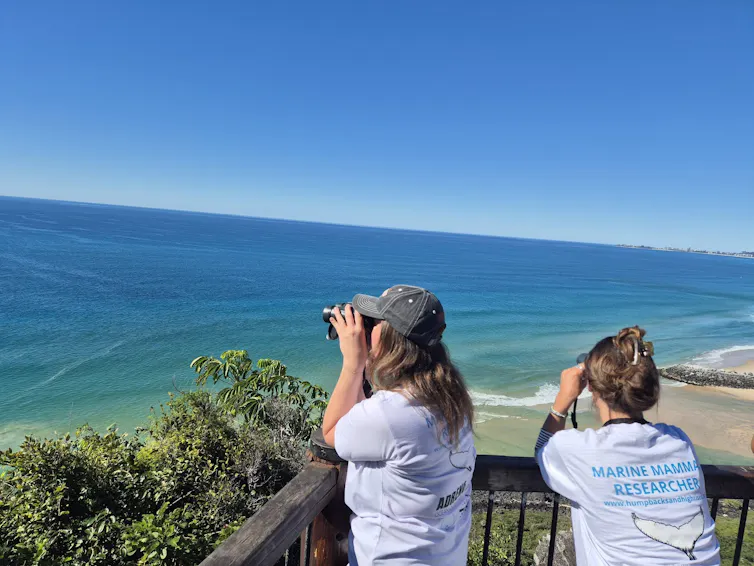 Two women holding binoculars look out at the ocean on a sunny day.