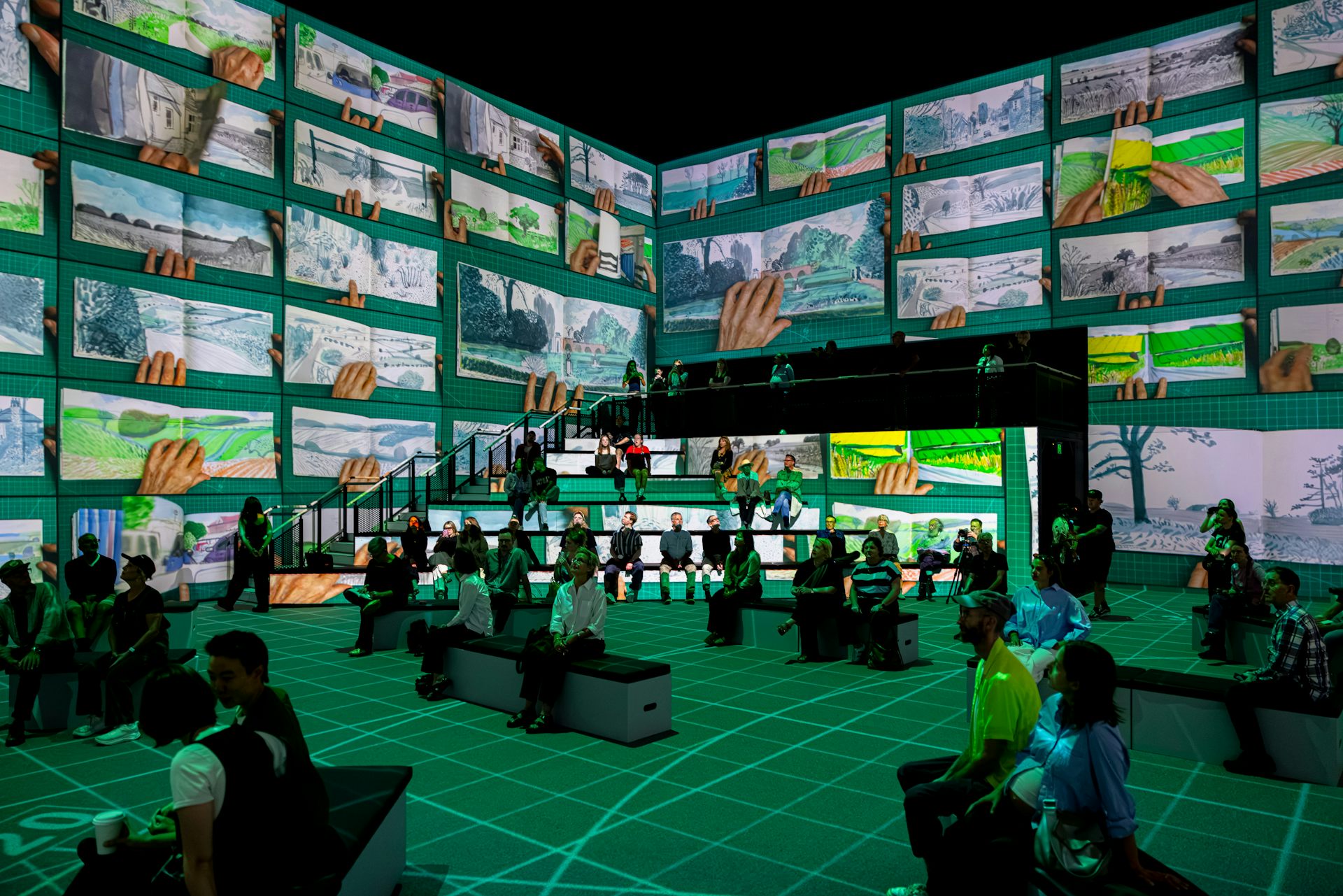 A darkened room with projections on the walls showing repeated birds-eye views of an artist sketchbook with artist's hands at work.