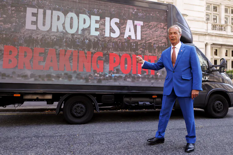 Small wrinkled man in an ill-fitting bright-blue suit standing before a bliboard truck reading 'Europe is at breaking point'.