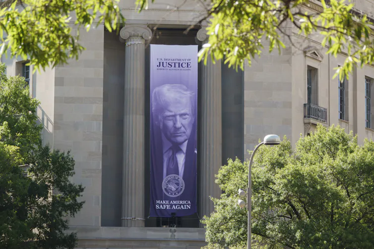 A long banner showing Donald Trump's face hangs between two grey pillars of a large building, which is partially obscured by trees with green leaves.