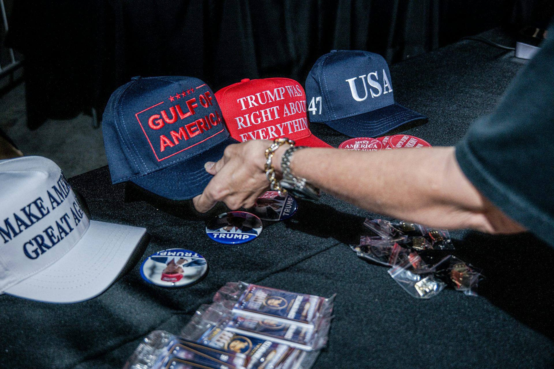 A person reaches out to pick up a Trump baseball cap with the words Gulf of America. Other caps have different messages.