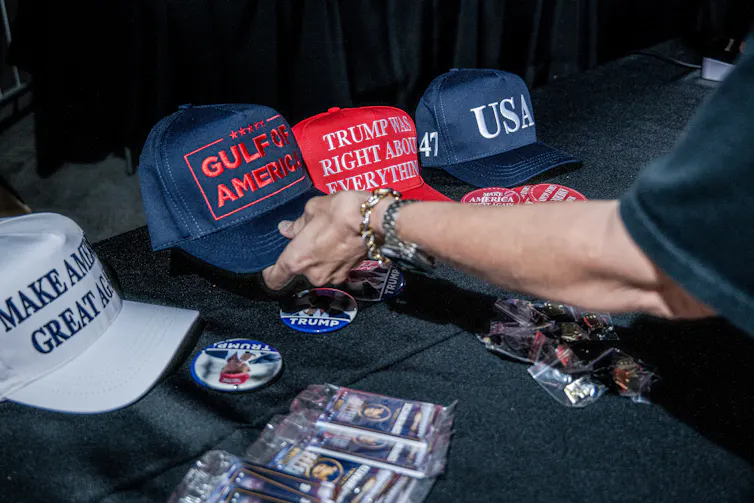 A person reaches out to pick up a Trump baseball cap with the words Gulf of America. Other caps have different messages.