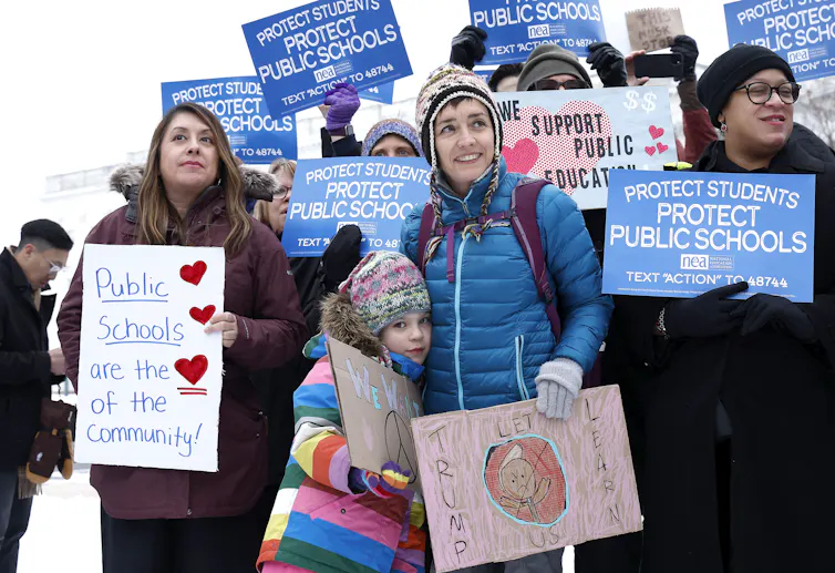 A group of people hold blue and white and other colorful signs that say 'Protect students, protect public schools.'