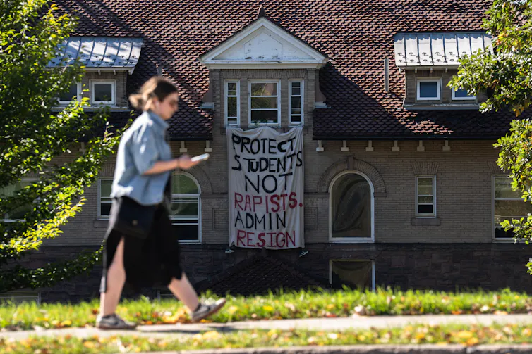 A woman looks at her phone and walks past a building that has a large cloth sign hanging off of it that says, 'Protect students, not rapists.'