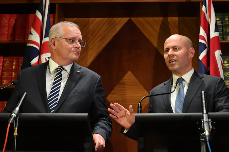 Two besuited men standing at adjacent podiums, one listening as the other speaks.